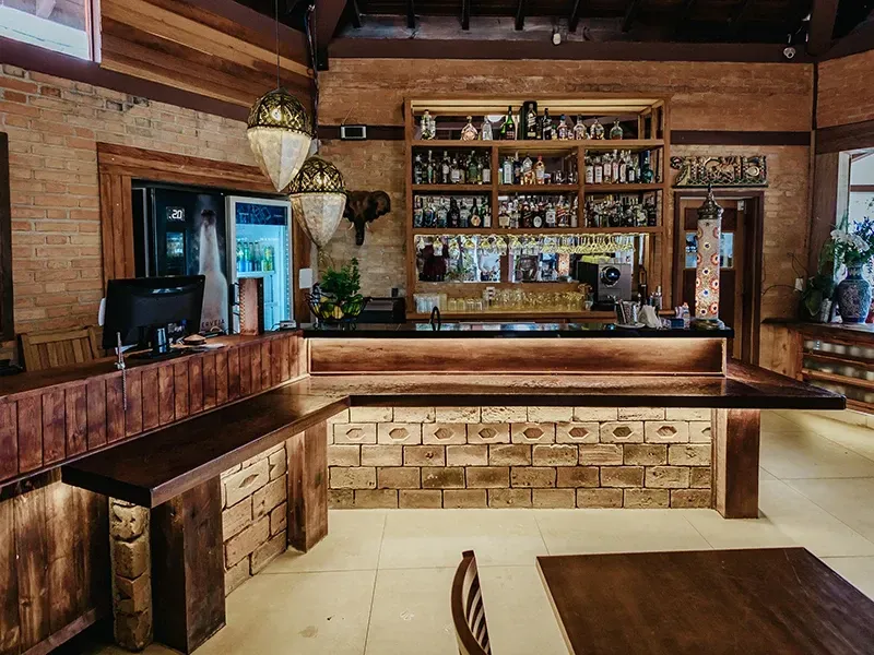 Um bar rústico com balcão de madeira, base de tijolos e prateleiras repletas de diversas garrafas, iluminado por luminárias pendentes.