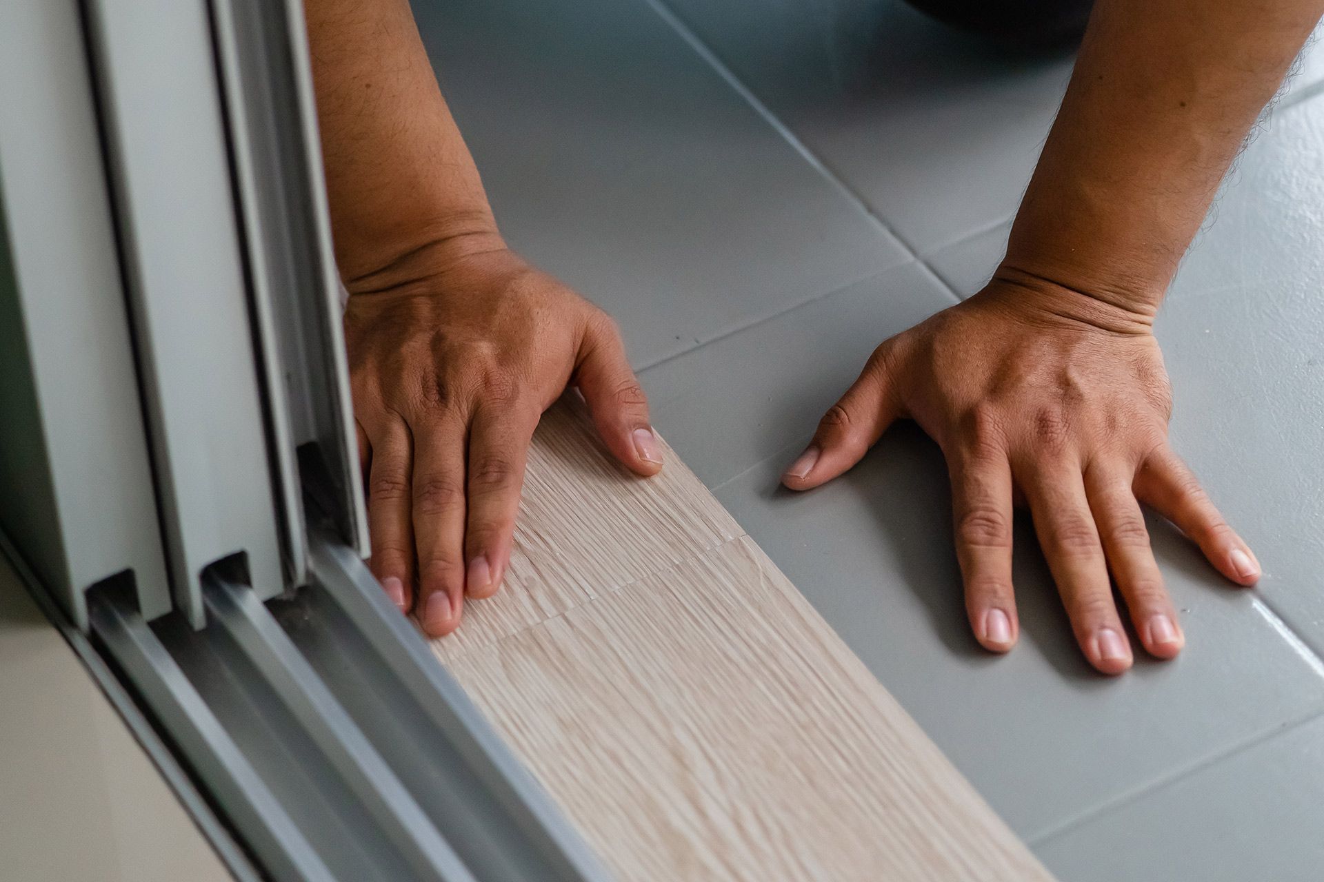 Hands measuring a floor transition strip beside a sliding door on gray tile flooring