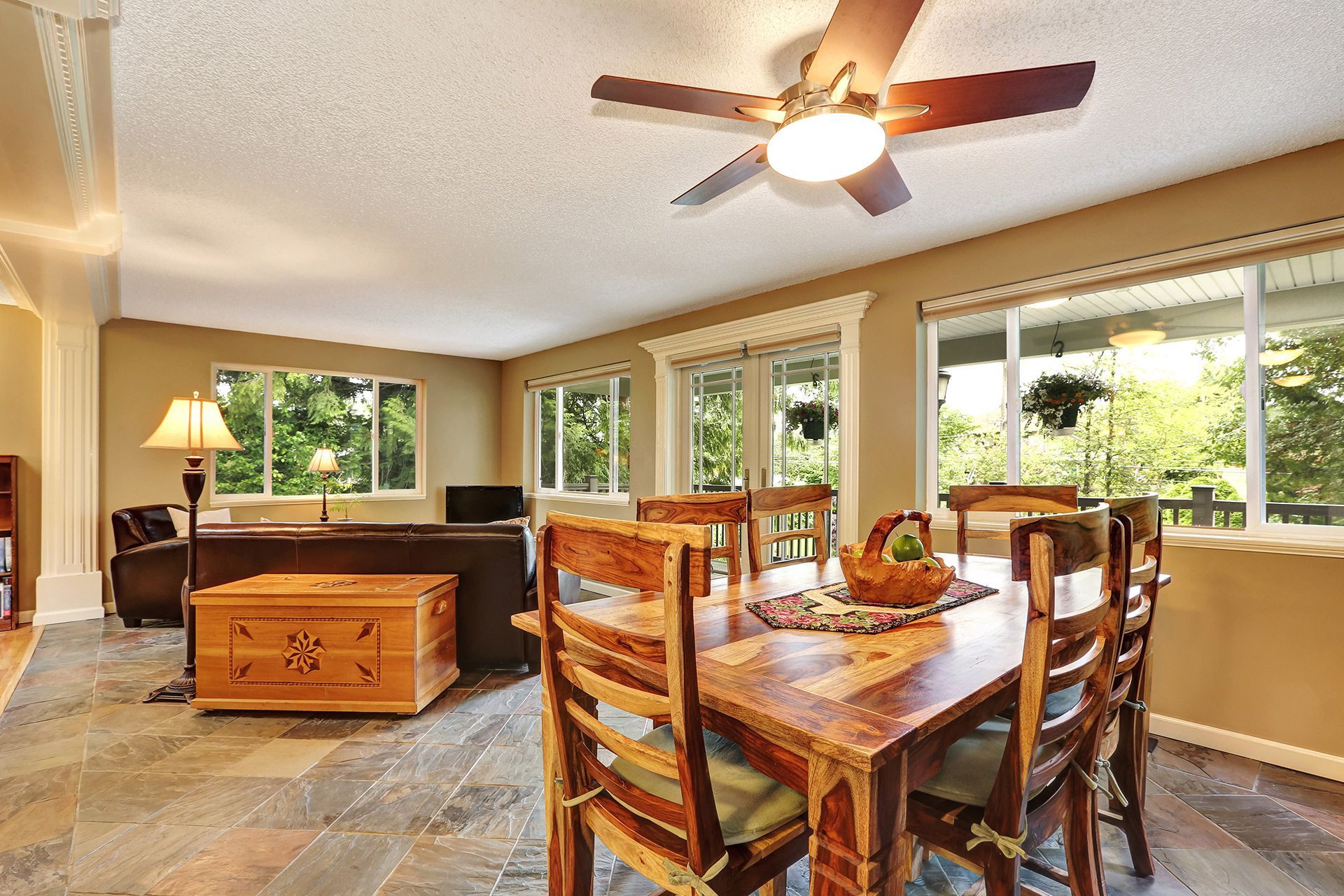 Open-concept living and dining room with large windows, wooden table, sectional sofa, and ceiling fan