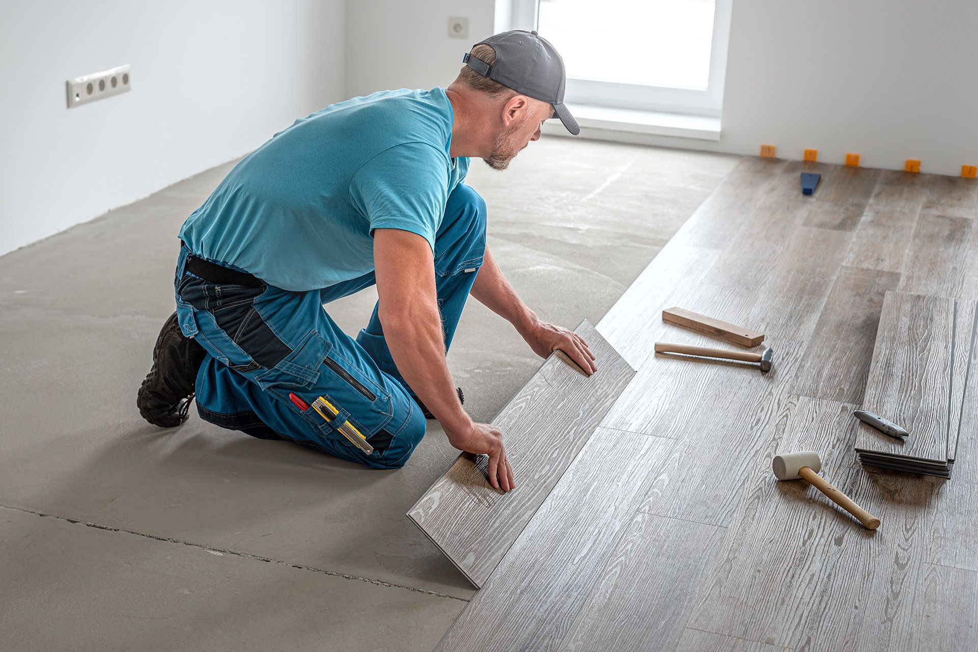 Worker kneeling on a floor, installing gray wood-look tiles in a bright room.