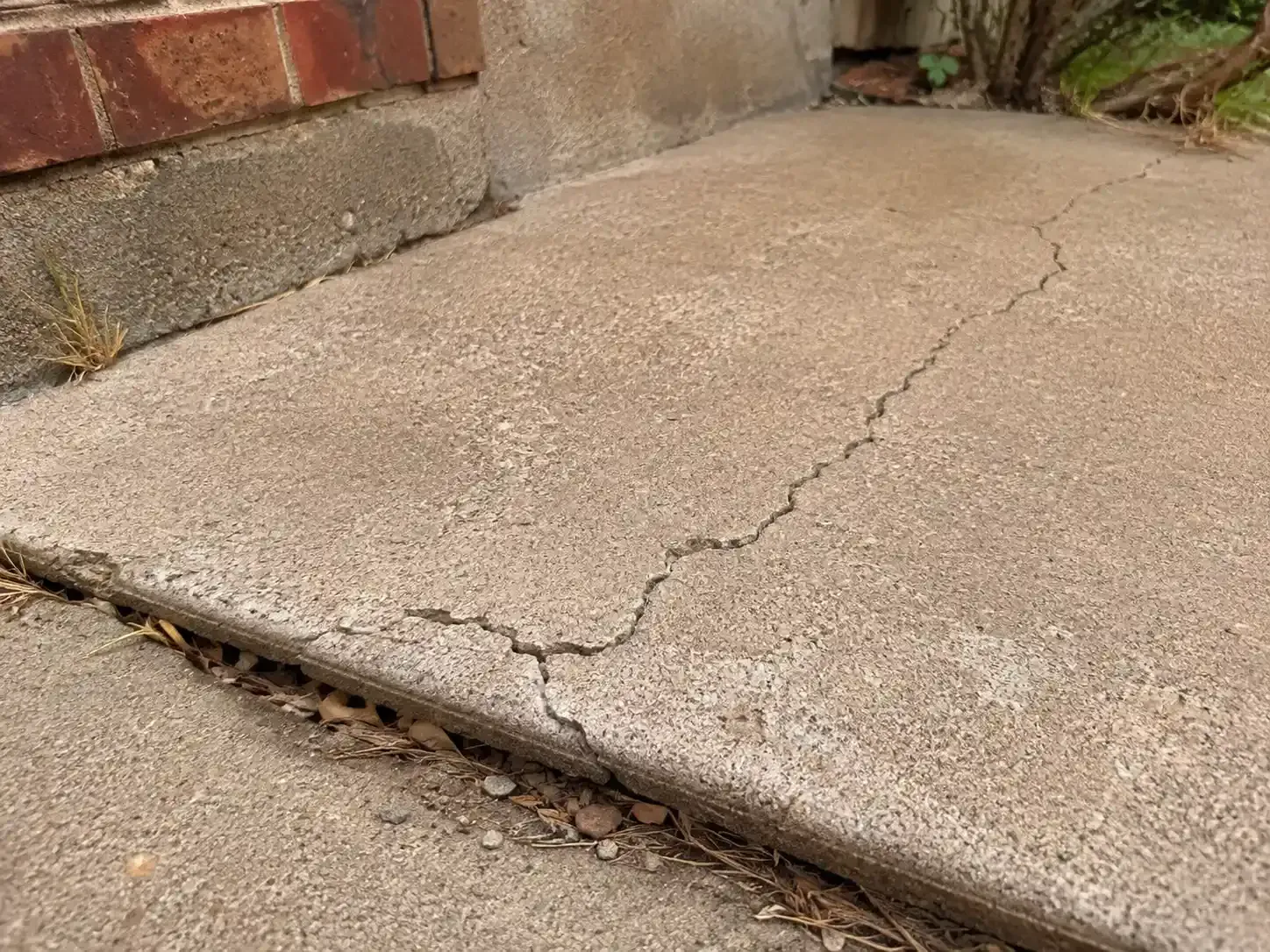 Cracked concrete patio slab with a raised edge beside a wall and gravel-lined gap