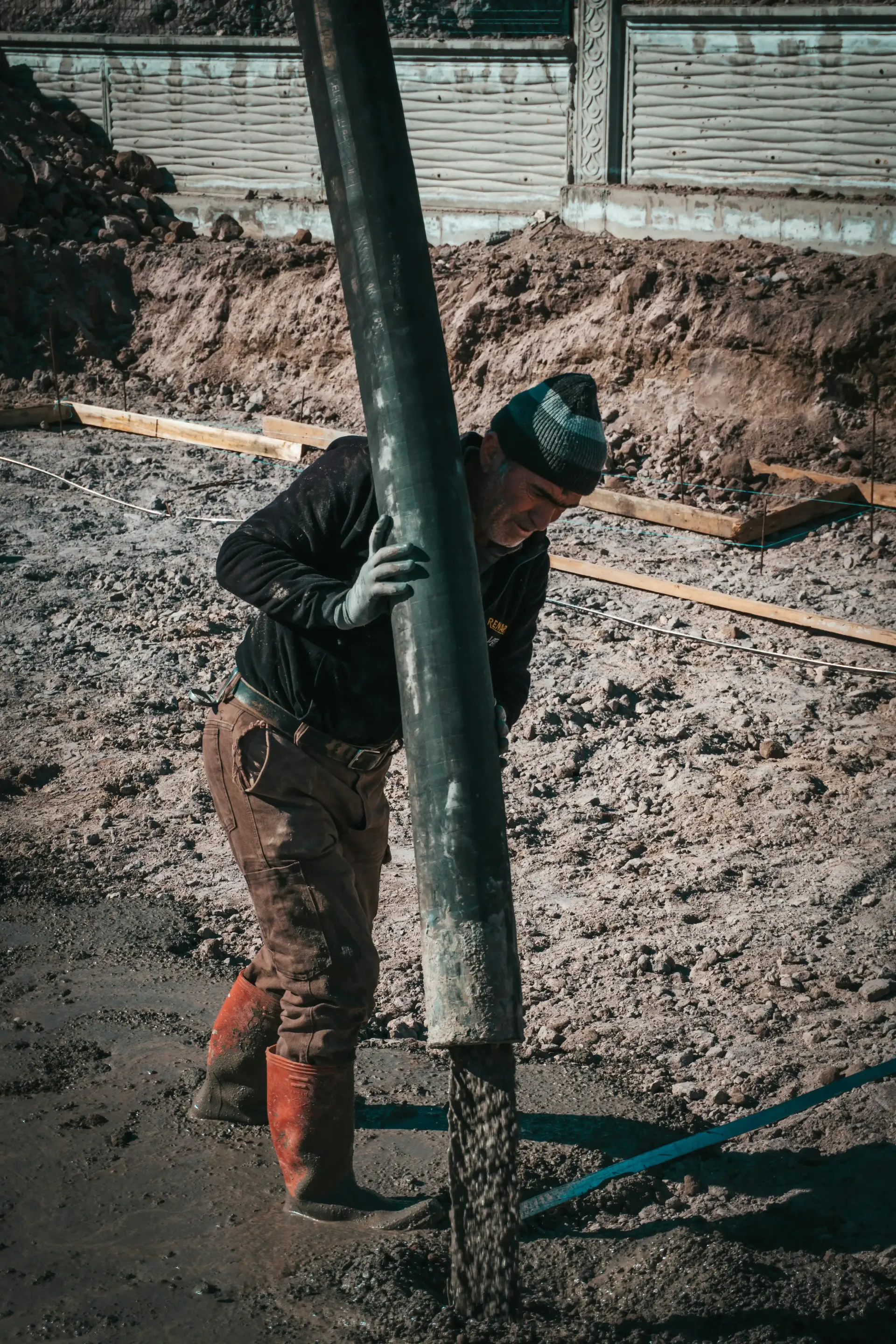 Worker holding concrete pouring chute pouring concrete foundation, wearing a dark jacket and boots.