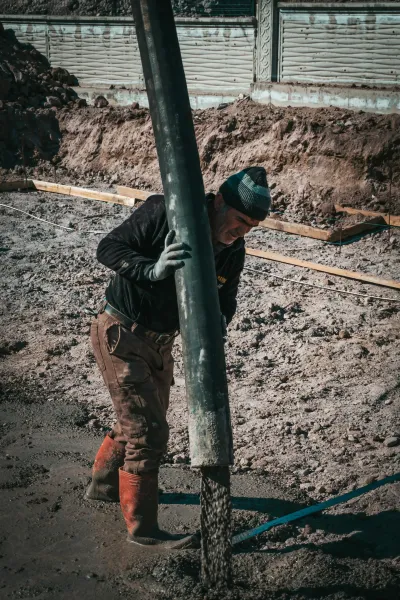 Worker holding concrete pouring chute pouring concrete foundation, wearing a dark jacket and boots.