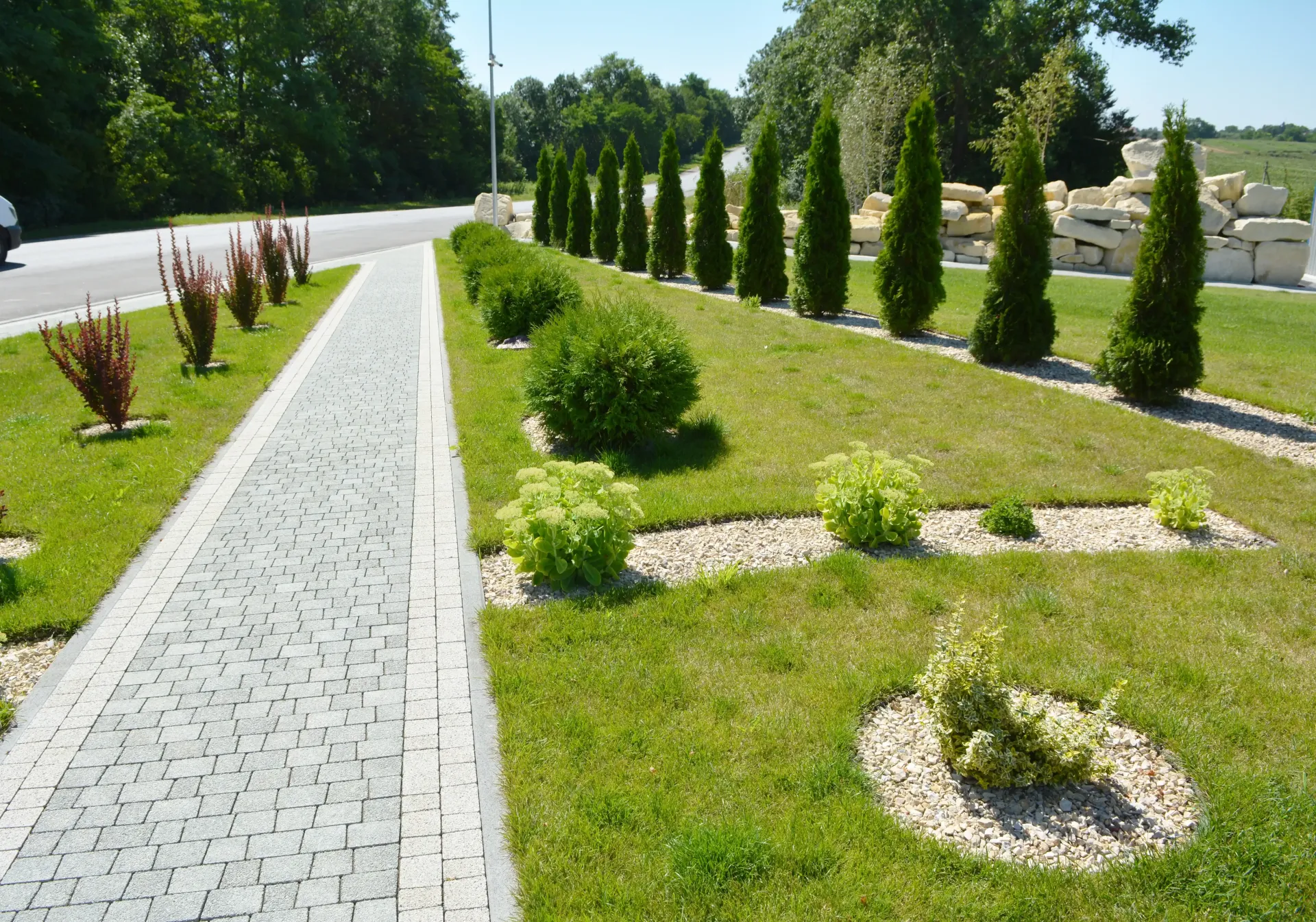Bright sun shining on concrete paver sidewalk , grass, and trimmed shrubs beside a road