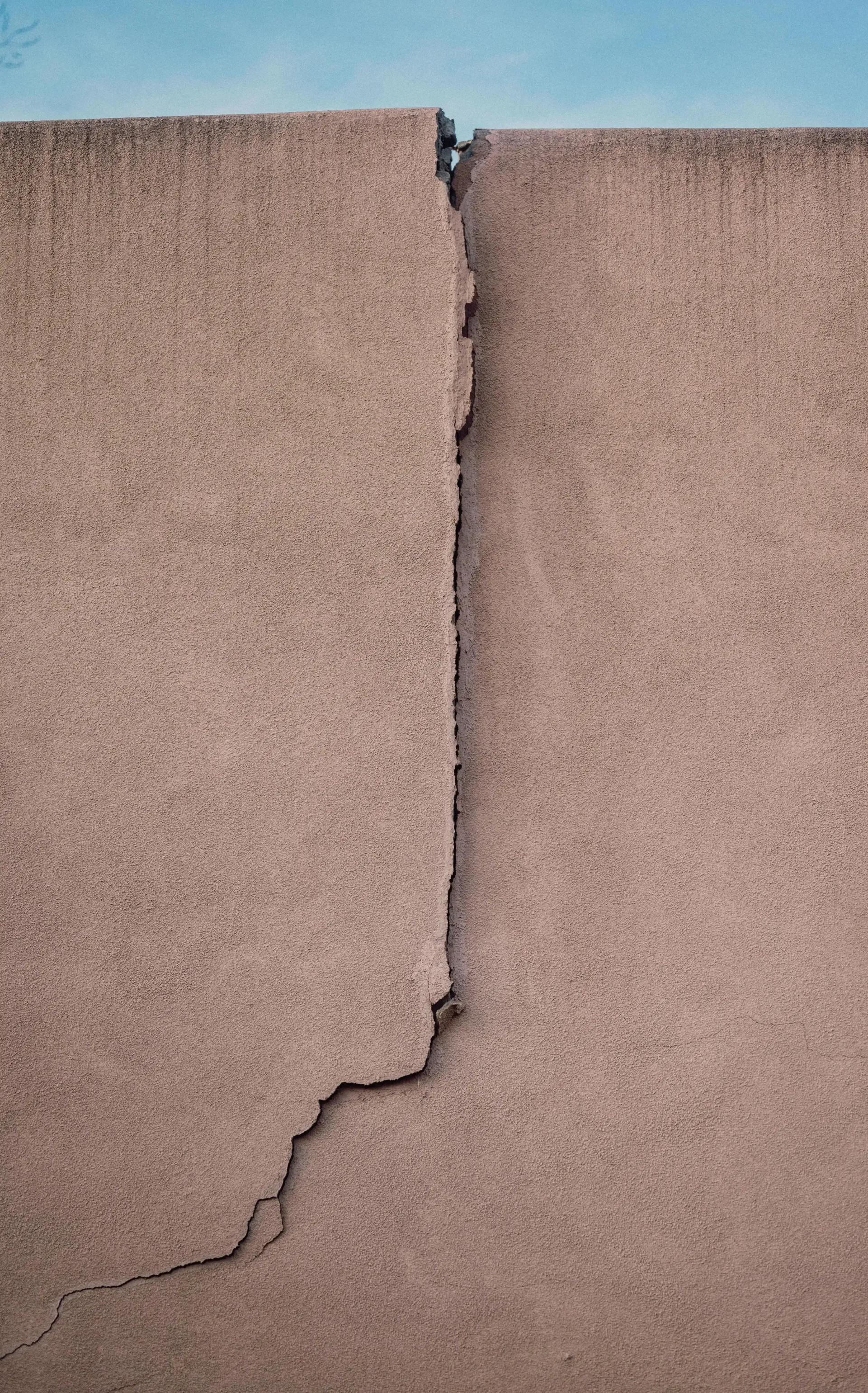 Cracked beige wall with a vertical and diagonal fissure under a blue sky