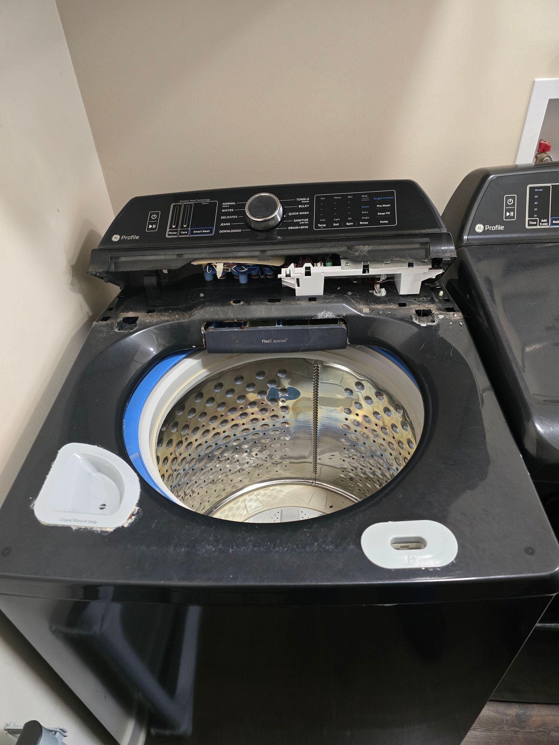 Black top-load washing machine with the lid open, revealing the drum. Another appliance is to the right.