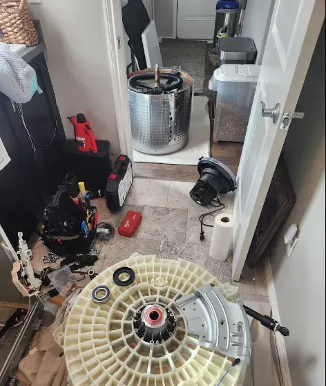 Washing machine disassembled in a small room. Parts are strewn on the floor, including the drum and motor.