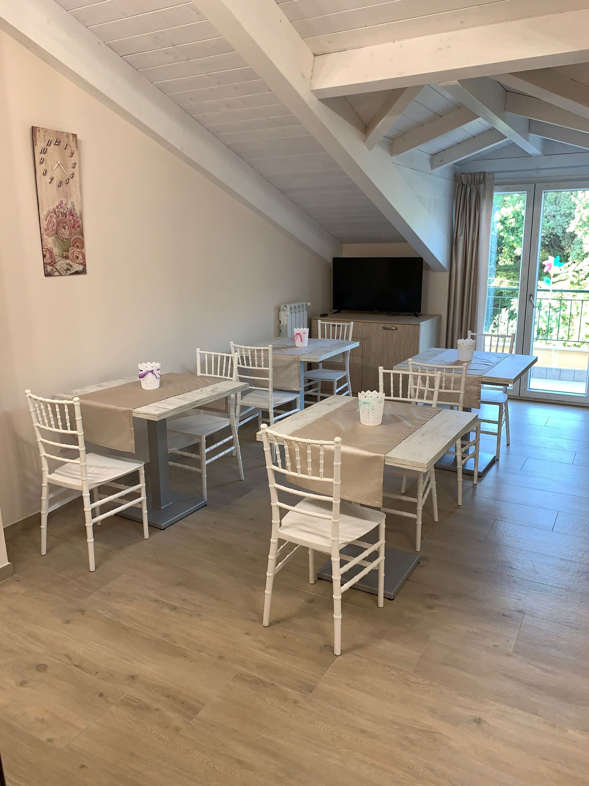 Sala da pranzo luminosa con sedie bianche, tavoli apparecchiati, TV e ampie finestre che si affacciano sul verde.