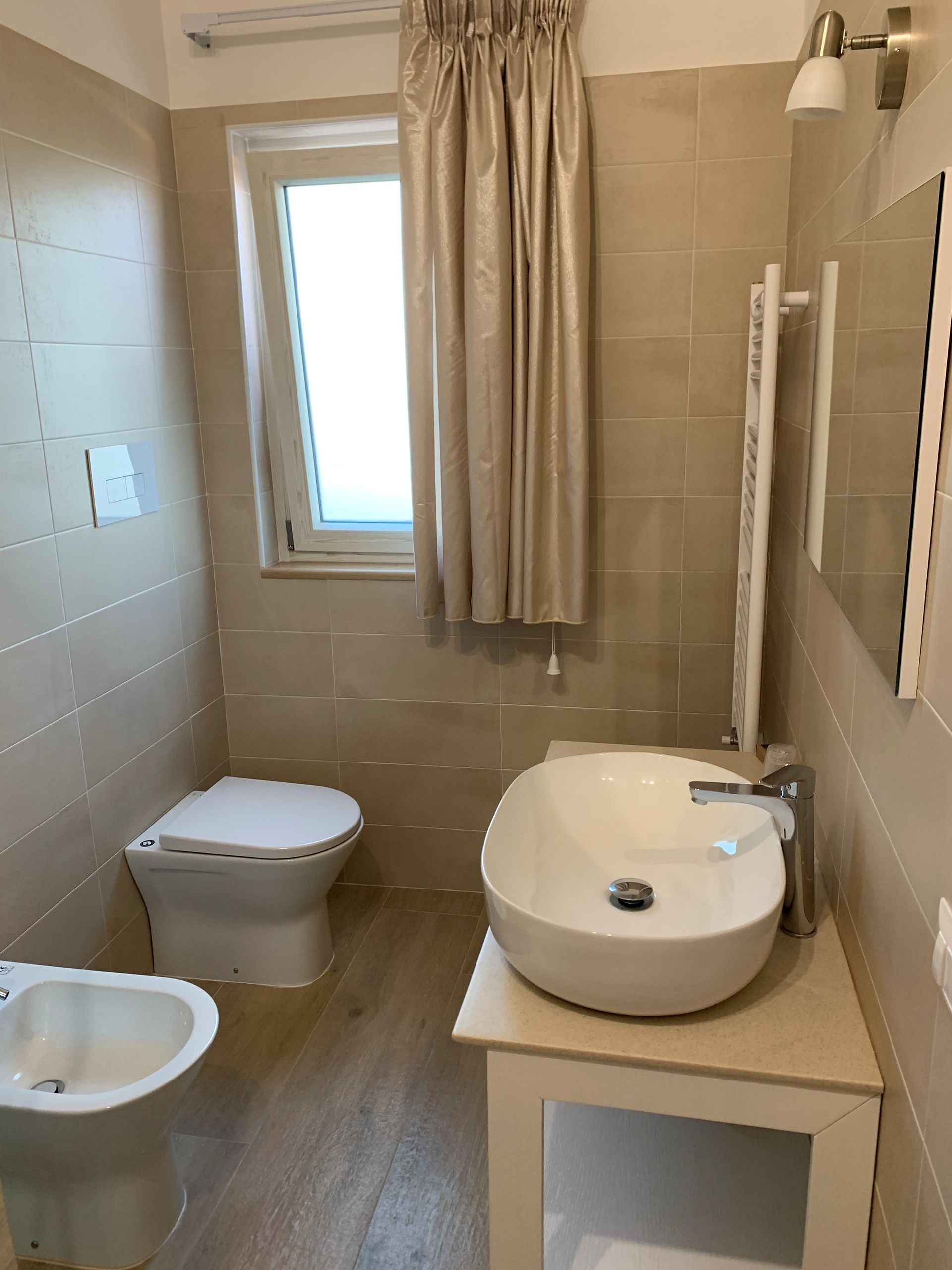 Piccolo bagno con WC, bidet, lavandino, specchio e finestra con tenda.