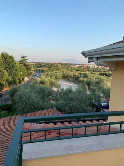 Vista dal balcone su un giardino rigoglioso, tetti di tegole rosse e colline in lontananza sotto un cielo azzurro e limpido.