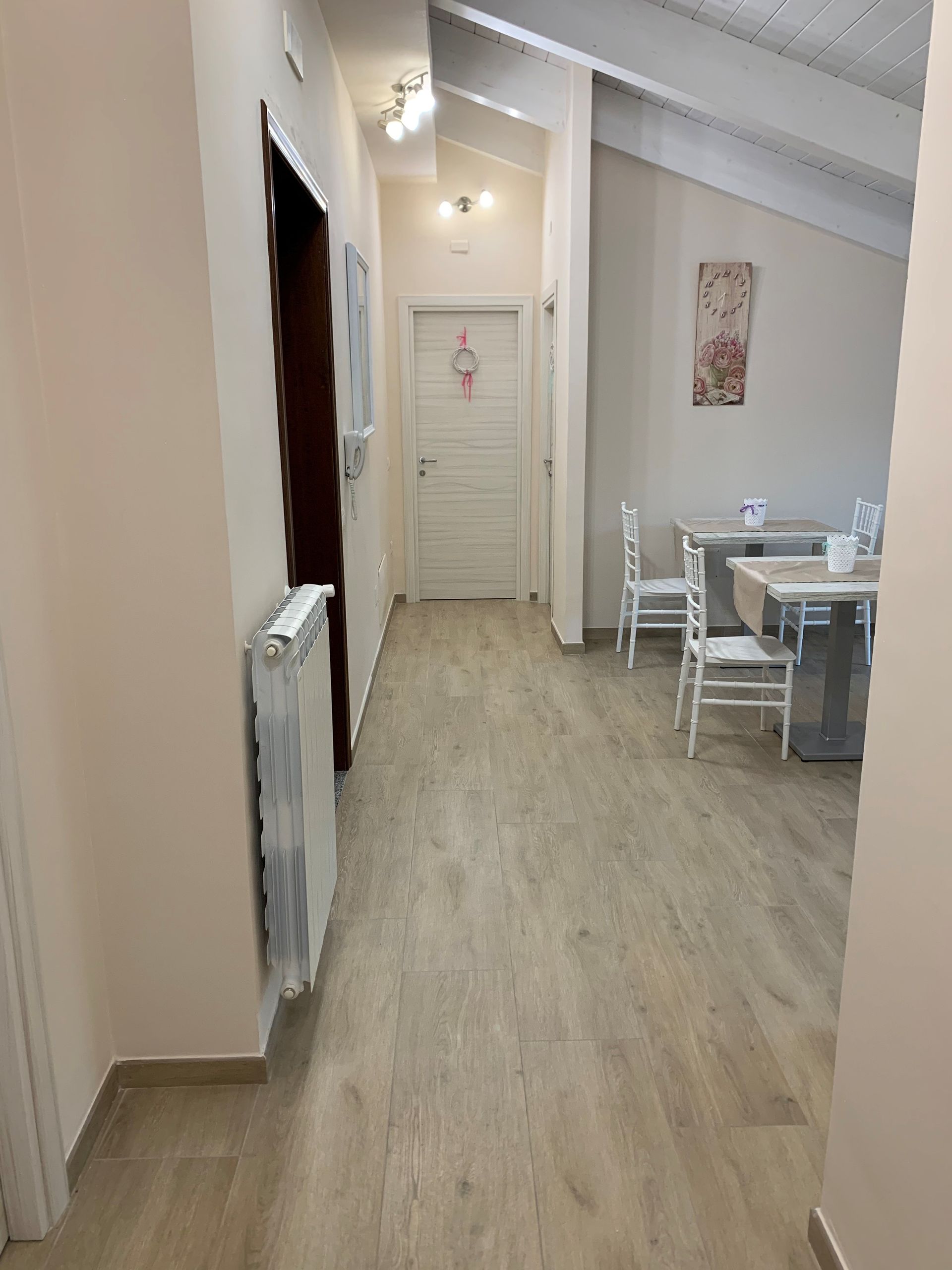 Stretto corridoio bianco con pavimento in legno, termosifone e, in fondo, un piccolo tavolo da pranzo con sedie.