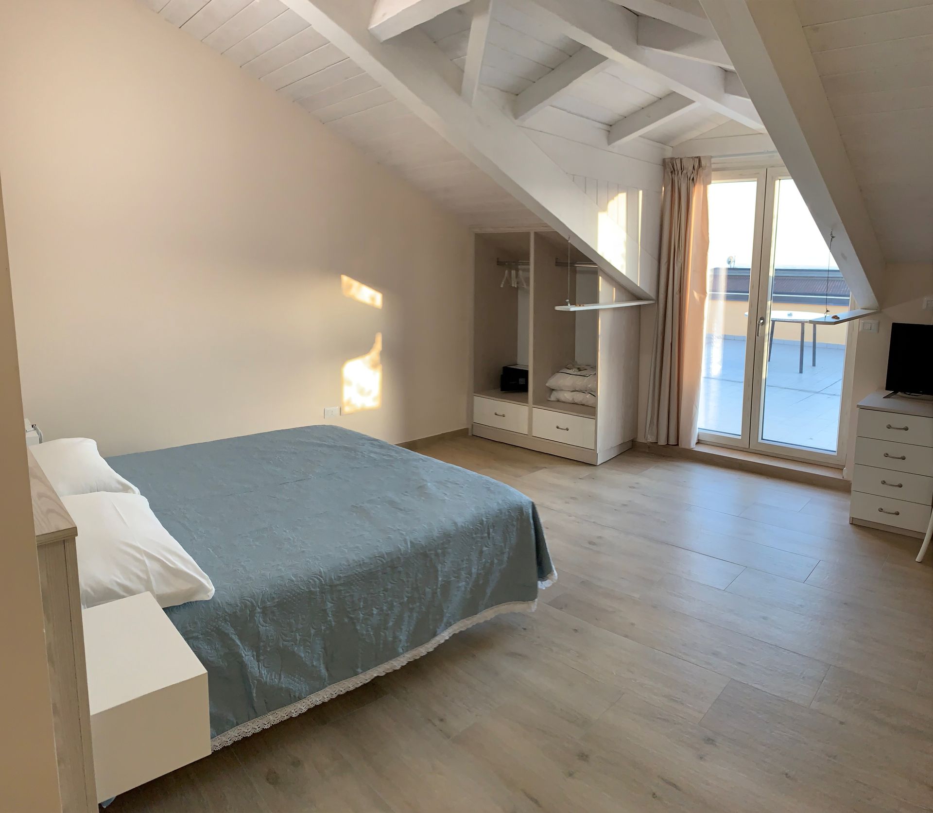 Luminosa camera da letto mansardata con soffitto inclinato, letto, comò e porte finestre che conducono a un balcone.