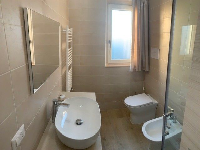 Luminoso bagno moderno con vasca, lavabo a parete, WC, specchio e finestra satinata.