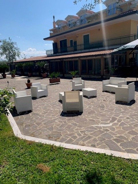 Patio esterno con sedie a sdraio bianche, pavimentazione in pietra e un edificio a due piani sotto un cielo azzurro brillante.