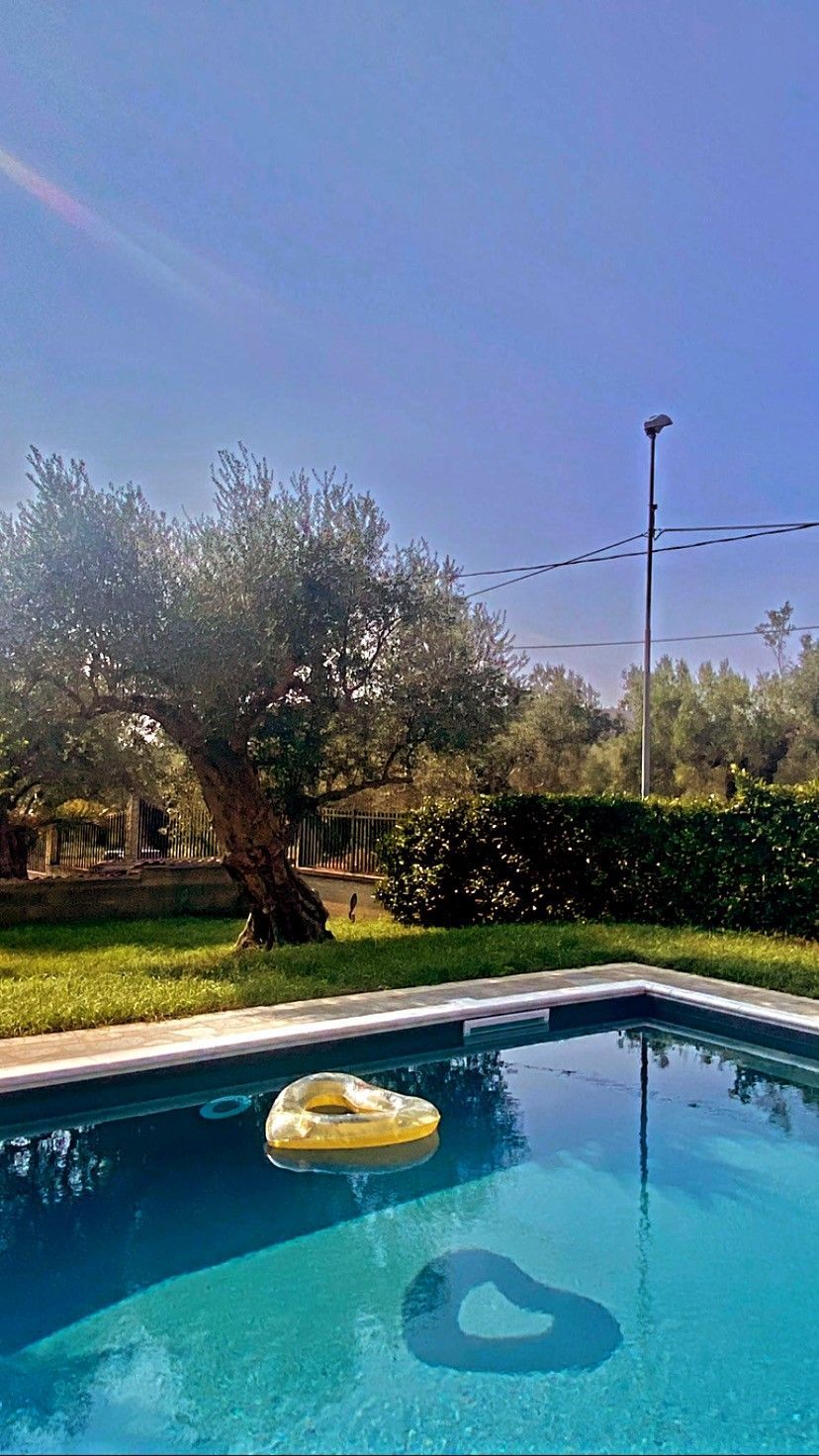 Piscina in giardino con un galleggiante giallo, alberi, siepe e un cielo azzurro limpido.
