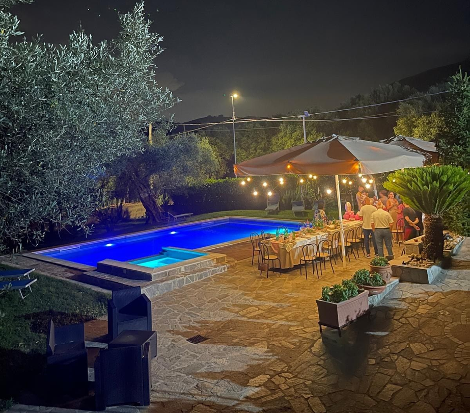 Terrazza notturna a bordo piscina con piscina illuminata di blu, luci decorative e persone che cenano sotto un grande ombrellone.