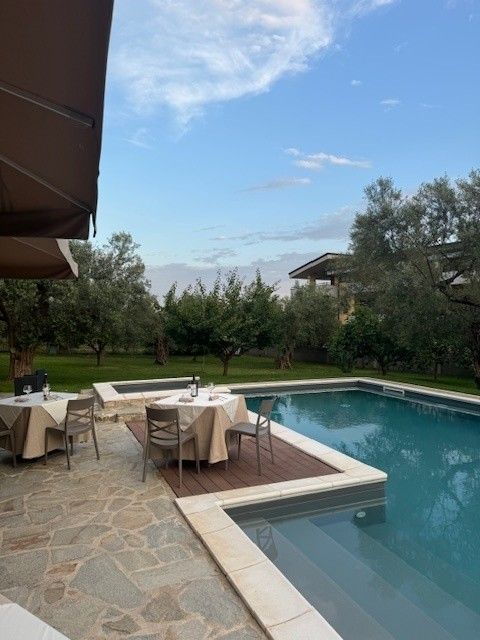 Patio esterno con tavoli e sedie a bordo piscina color turchese, sotto un cielo parzialmente nuvoloso.