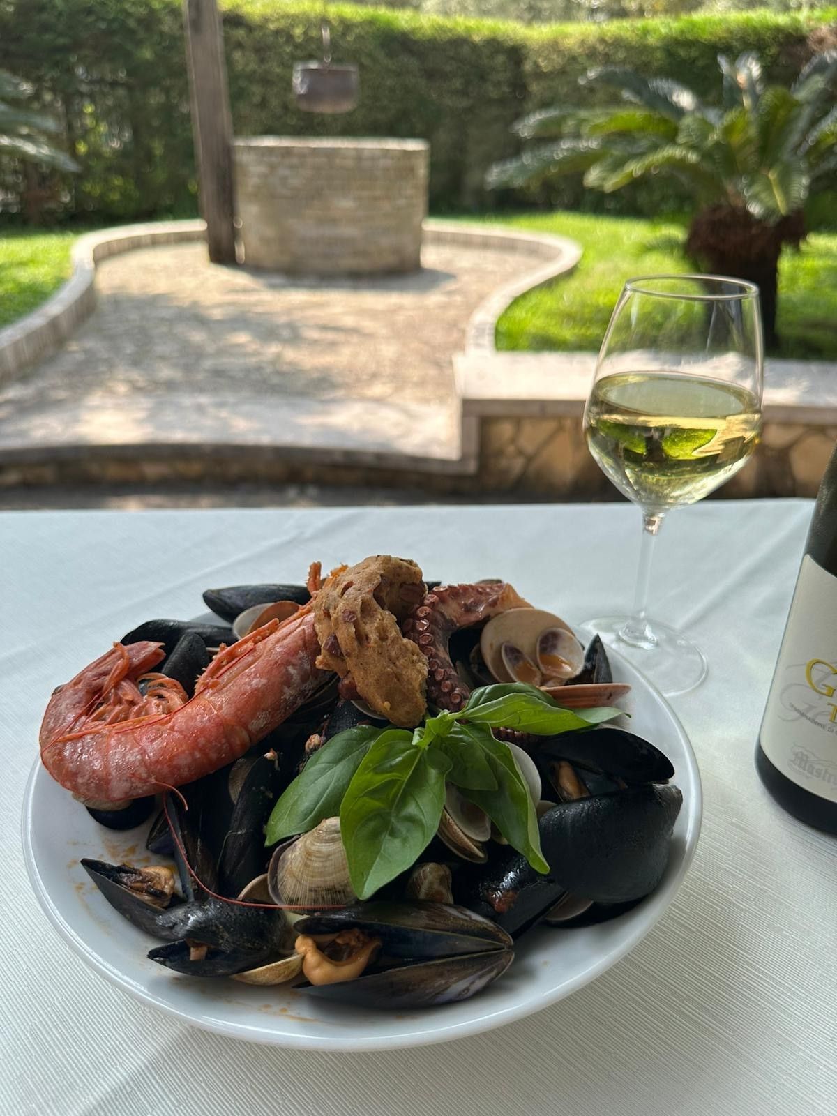 Pasta ai frutti di mare con cozze e gamberi su un tavolo accanto a un calice di vino, con vista su una terrazza con giardino.