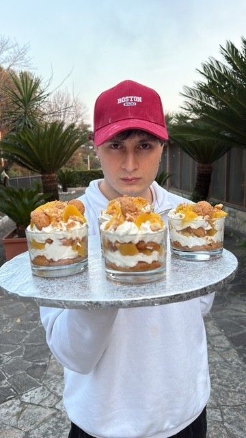 Una persona con un berretto rosso tiene in mano un vassoio di coppette di dessert a strati, guarnite con panna montata, all'aperto.