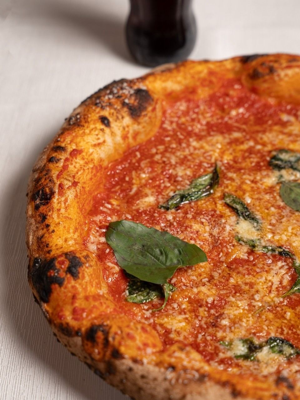 Primo piano di una pizza margherita con salsa di pomodoro, foglie di basilico e crosta bruciacchiata su una tovaglia bianca.