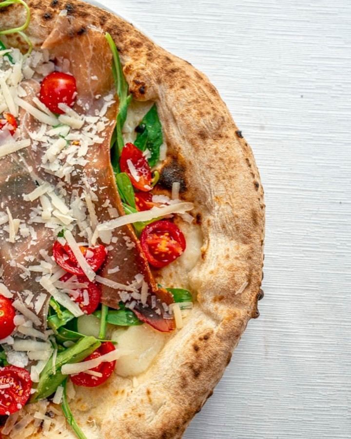 Primo piano di una pizza vegetariana con pomodorini, verdure a foglia verde, formaggio e funghi su una superficie bianca.