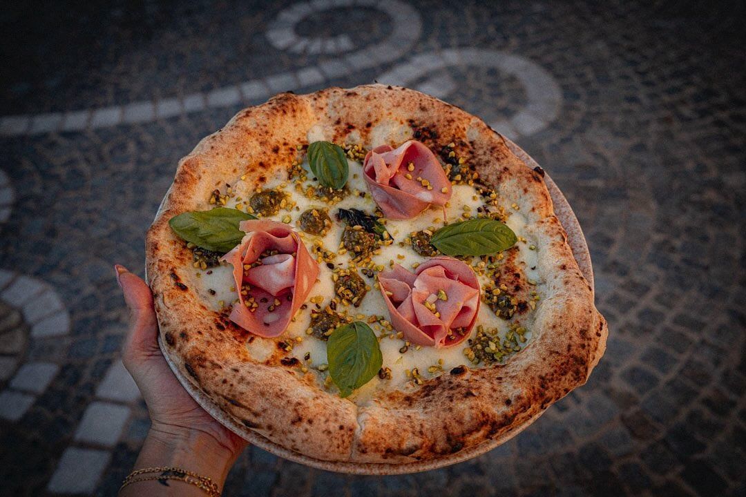 Pizza napoletana con prosciutto, peperoni verdi e basilico su un piatto rotondo sopra una tovaglia decorata.