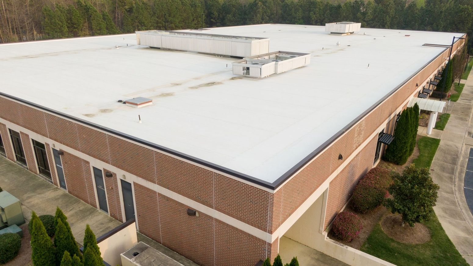 Aerial view of a brick industrial building with a white flat roof and surrounding trees