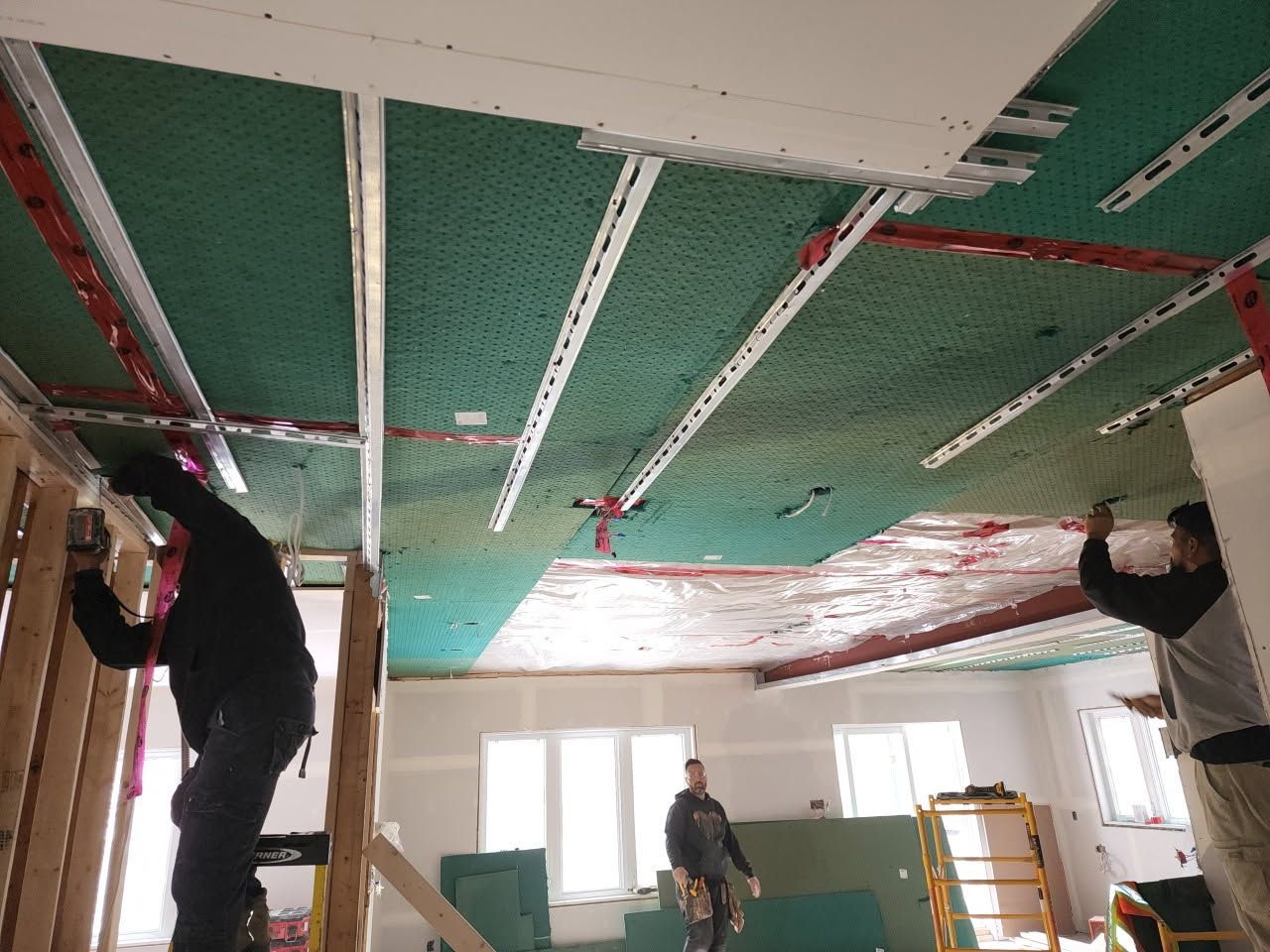 Des ouvriers du bâtiment installent des panneaux de plafond, laissant apparaître l'isolant vert. Intérieur avec outils et fenêtres.