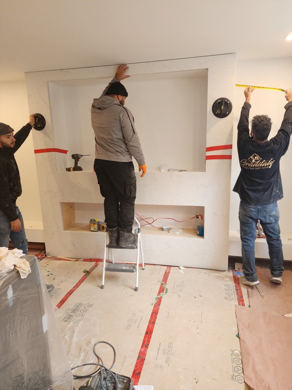 Trois personnes prennent des mesures et travaillent sur un élément mural encastré dans une pièce en construction.