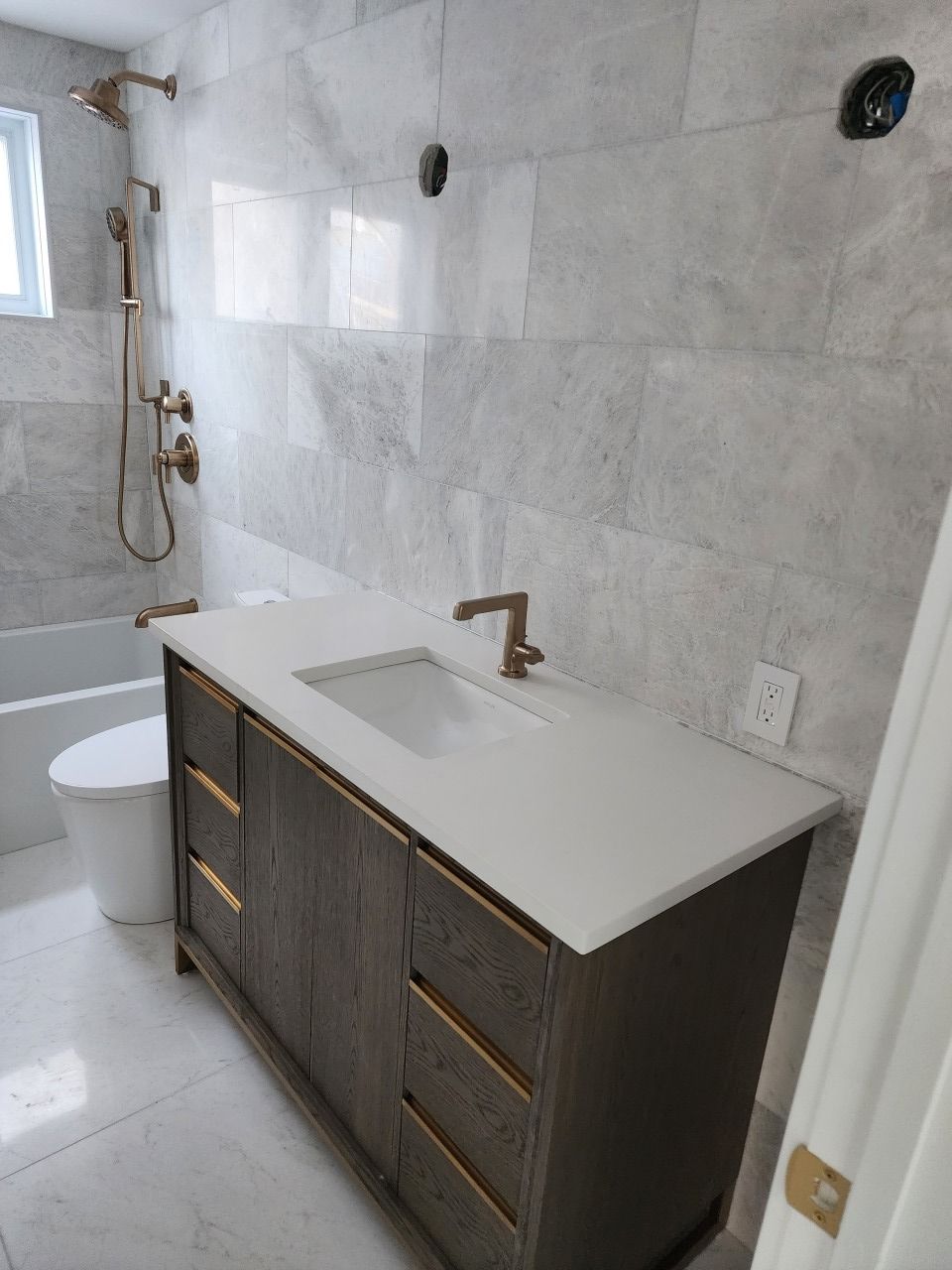 Salle de bains moderne avec murs carrelés gris, meuble-lavabo, lavabo, baignoire et robinetterie dorée.