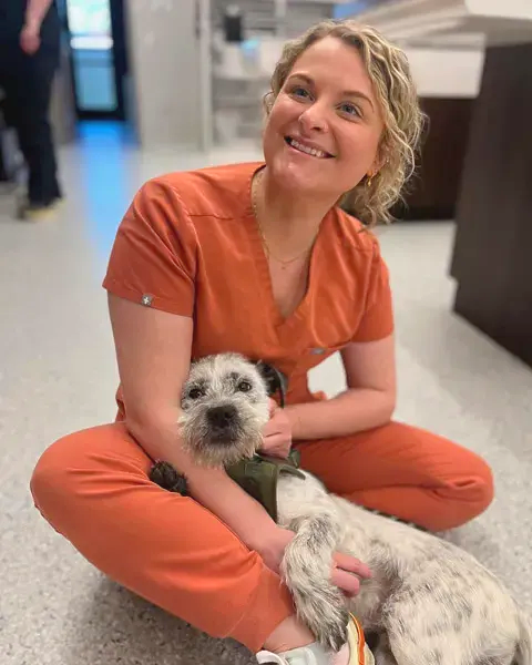 Smiling person in orange scrubs sits on the floor holding a small white-and-black dog in a hallway