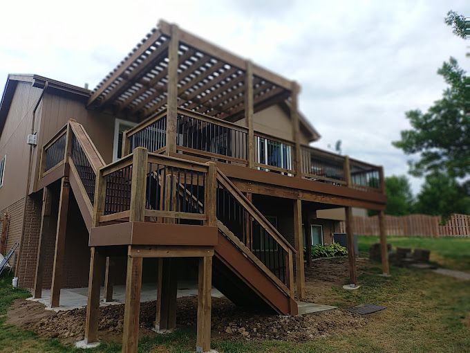 A two-story wooden deck with a built-in pergola and staircase, attached to the back of a brown house.