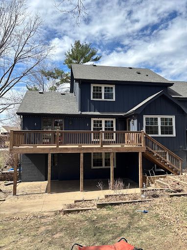 A two-story dark blue house with a wooden deck, a patio underneath, and a staircase leading down to a lawn.