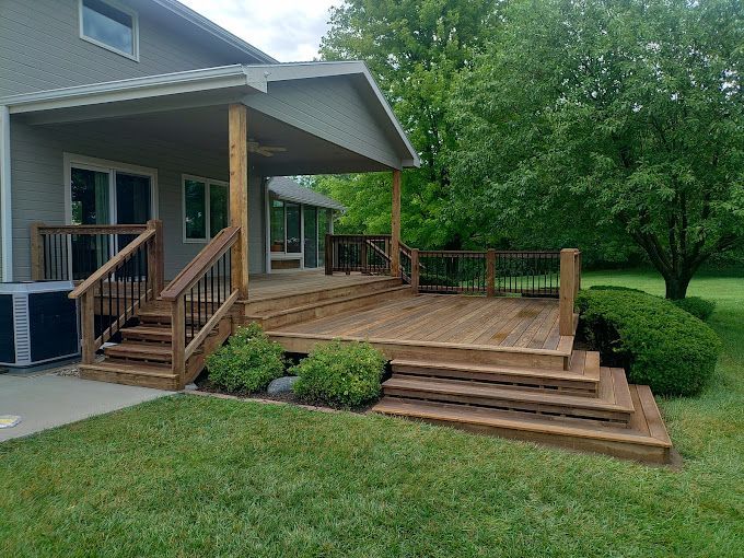A wooden deck with stairs and a covered patio area attached to a gray house with a lawn and trees in the background.