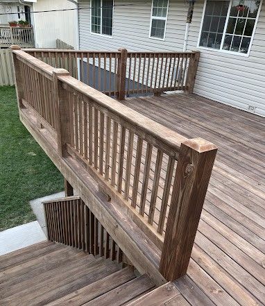 A wooden deck with a staircase leading down to a concrete path, featuring brown railings against a light-sided house.