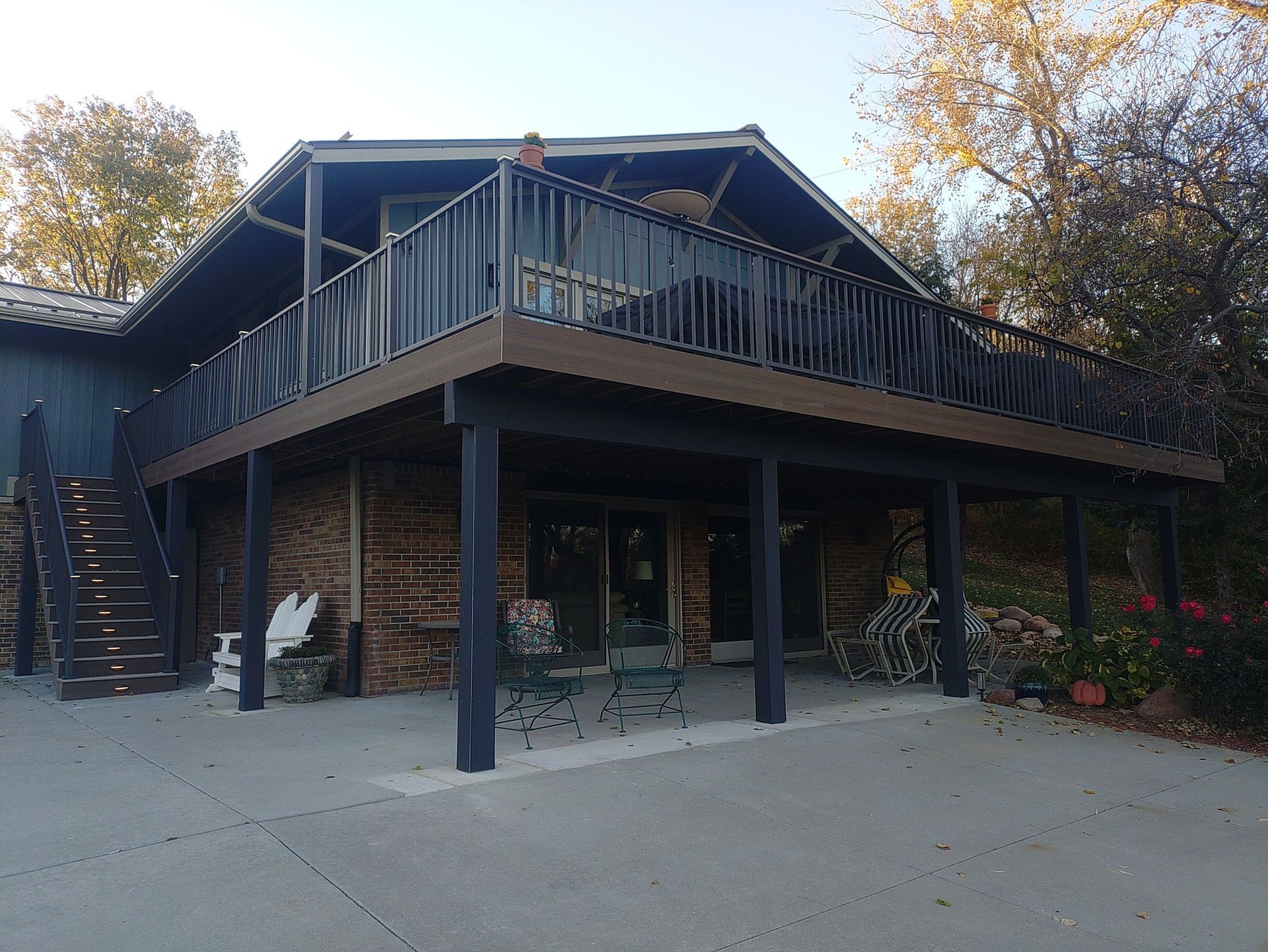 A two-story house with a large upper deck and black railings, overlooking a patio with stairs on the left side.