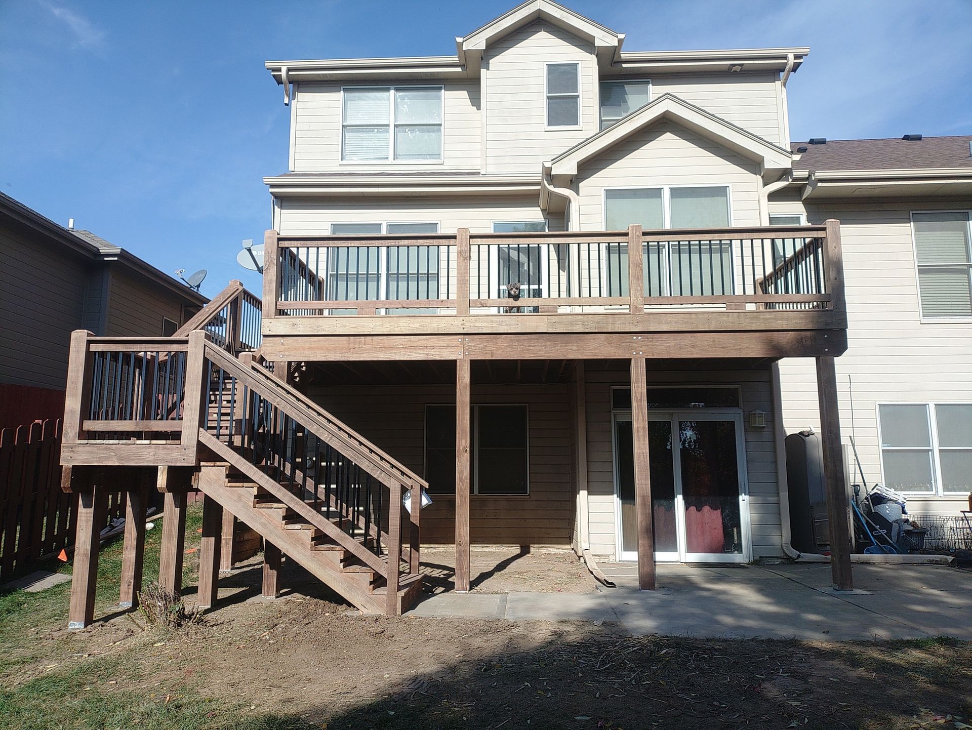 A beige two-story house with a wooden deck and staircase leading to a ground-level concrete patio.