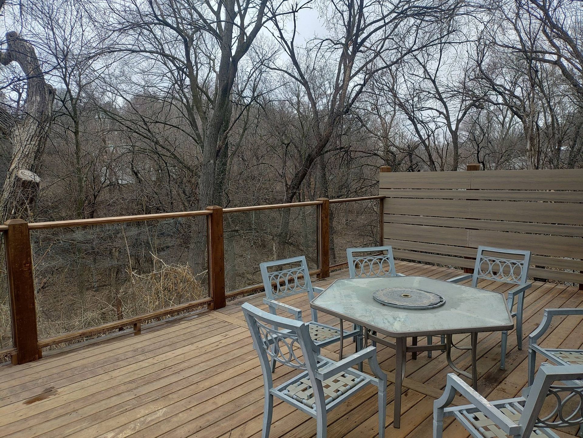 A wooden deck features a hexagonal table with five chairs, overlooking a backdrop of bare trees on a cloudy day.
