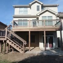 Back of a two-story beige house with a wooden deck and a set of stairs leading to a patio.