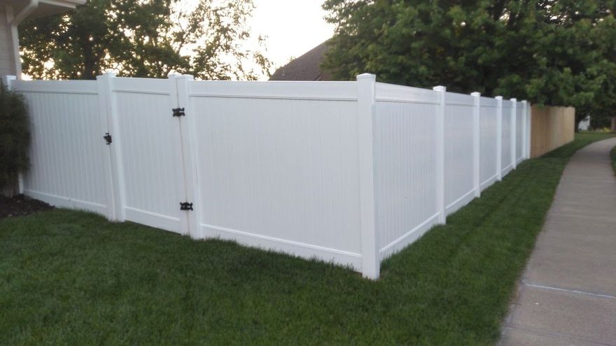 White vinyl fence enclosing a green lawn next to a sidewalk and a tree line.