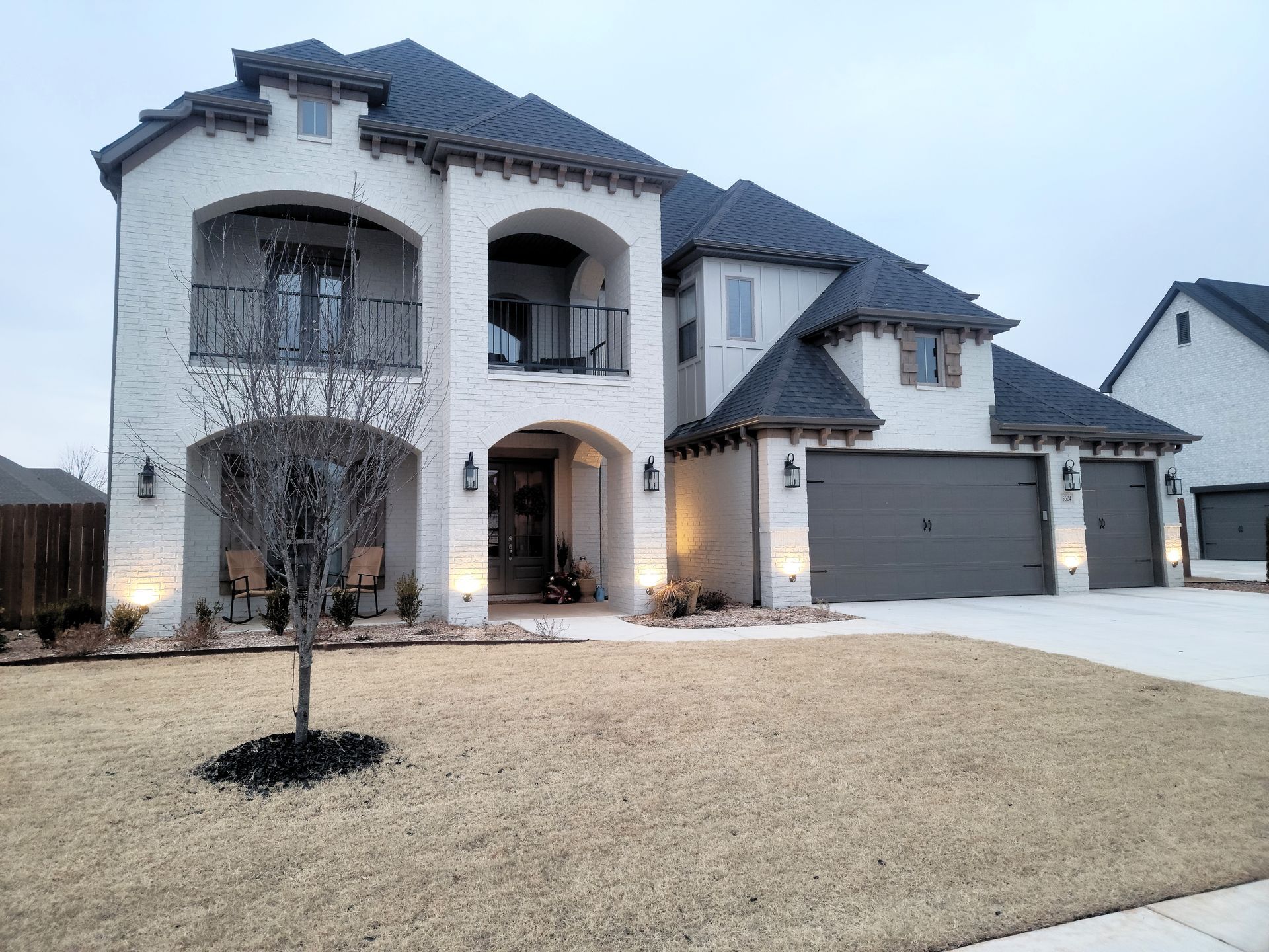 Large two-story stone house with a gray roof, arched entryways, and a wide driveway at dusk