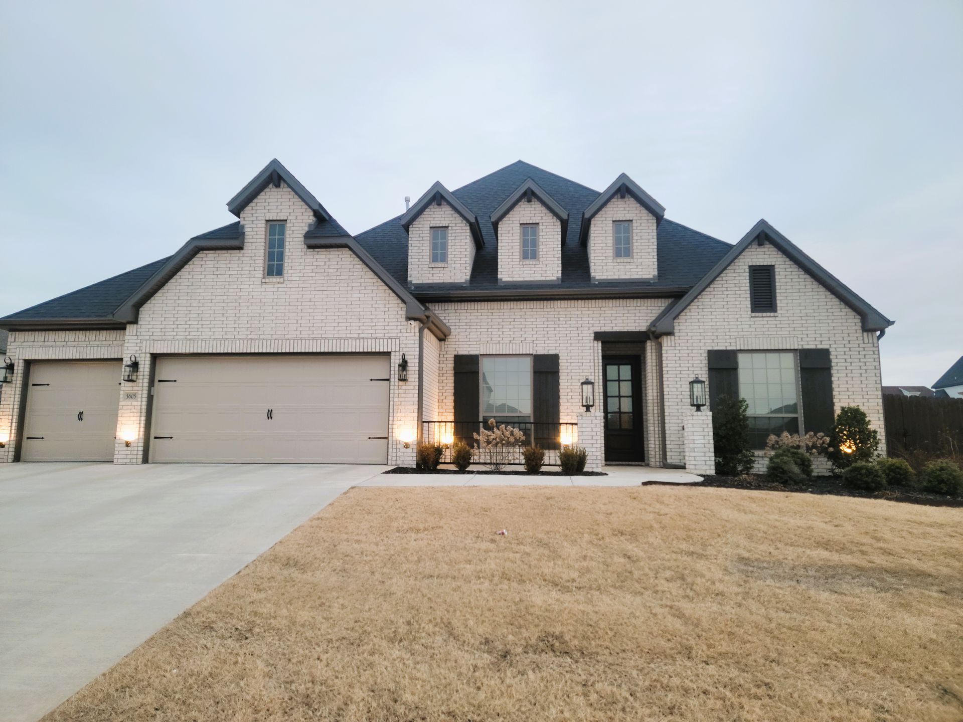 Suburban two-story house with a three-car garage, stone accents, and a lit front porch at dusk