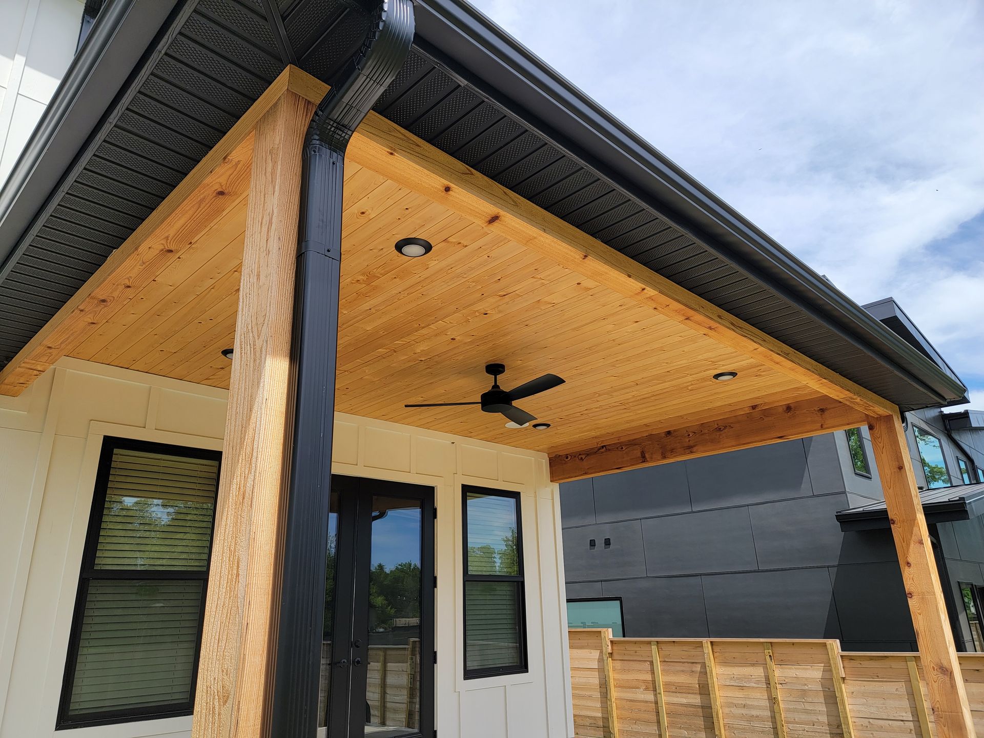 Modern porch with wood ceiling, black fan, white door, windows, and dark exterior siding.
