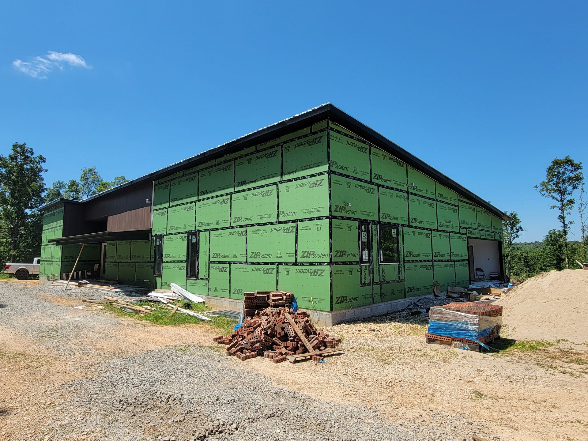 Green barn-like building under construction on a gravel lot, with lumber and sand piles outside.