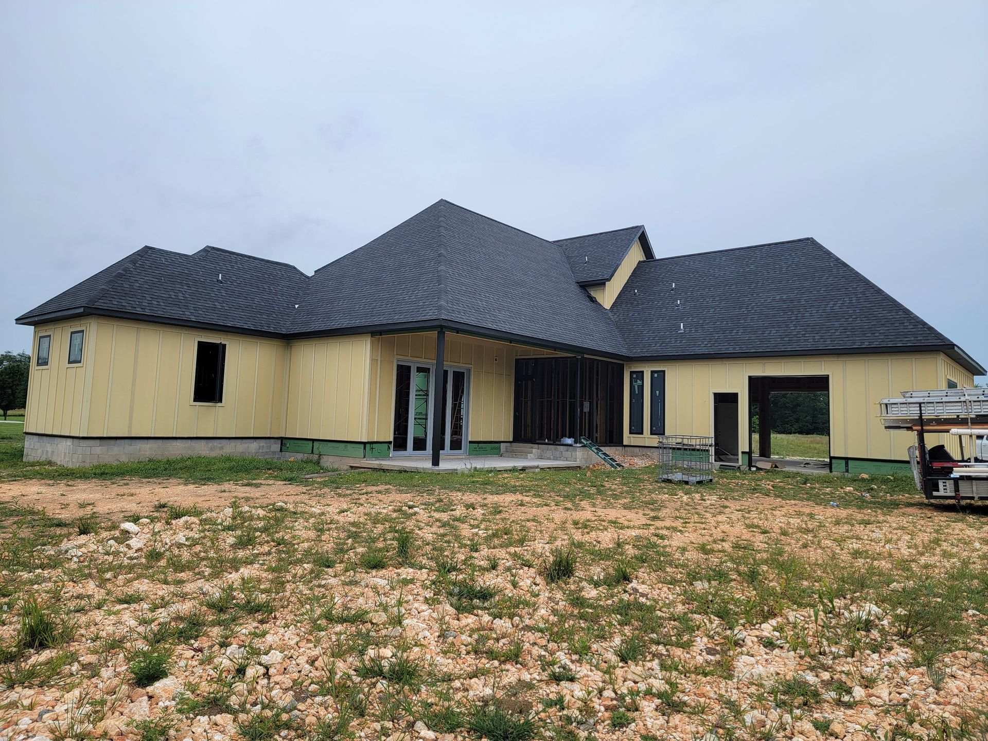 Single-story house under construction with dark roof and unfinished yard on an overcast day