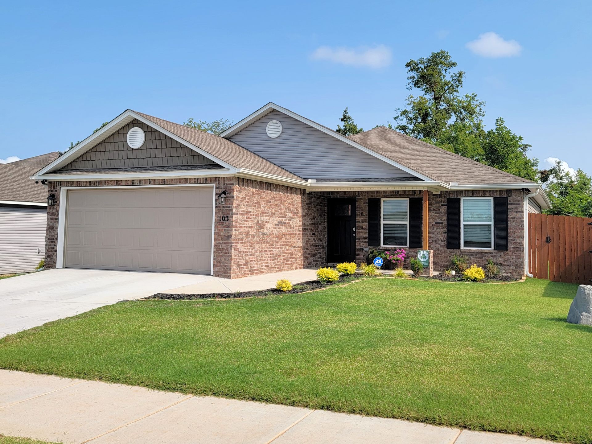 Single-story brick house with two-car garage, manicured lawn, and flower beds under a blue sky