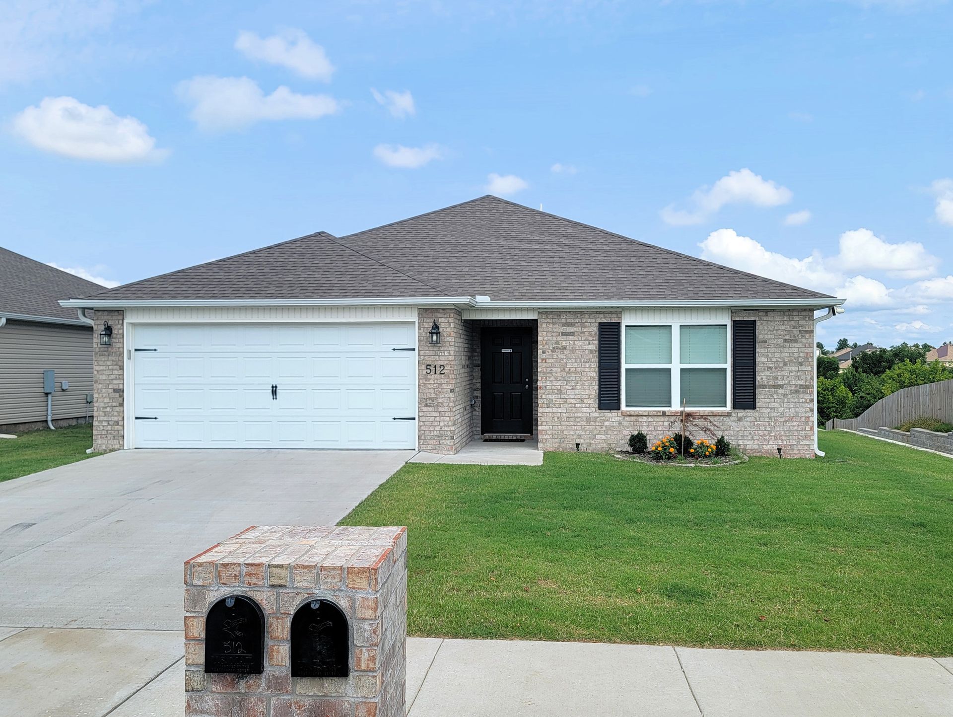 Single-story brick house with a two-car garage, front lawn, and mailbox under a blue sky.