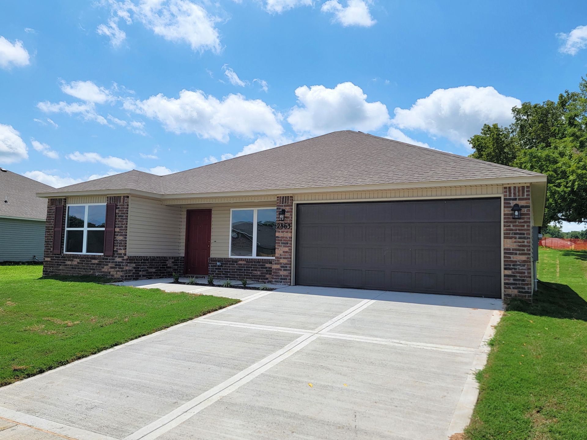 Single-story brick house with a two-car garage, concrete driveway, and green lawn under a blue sky.