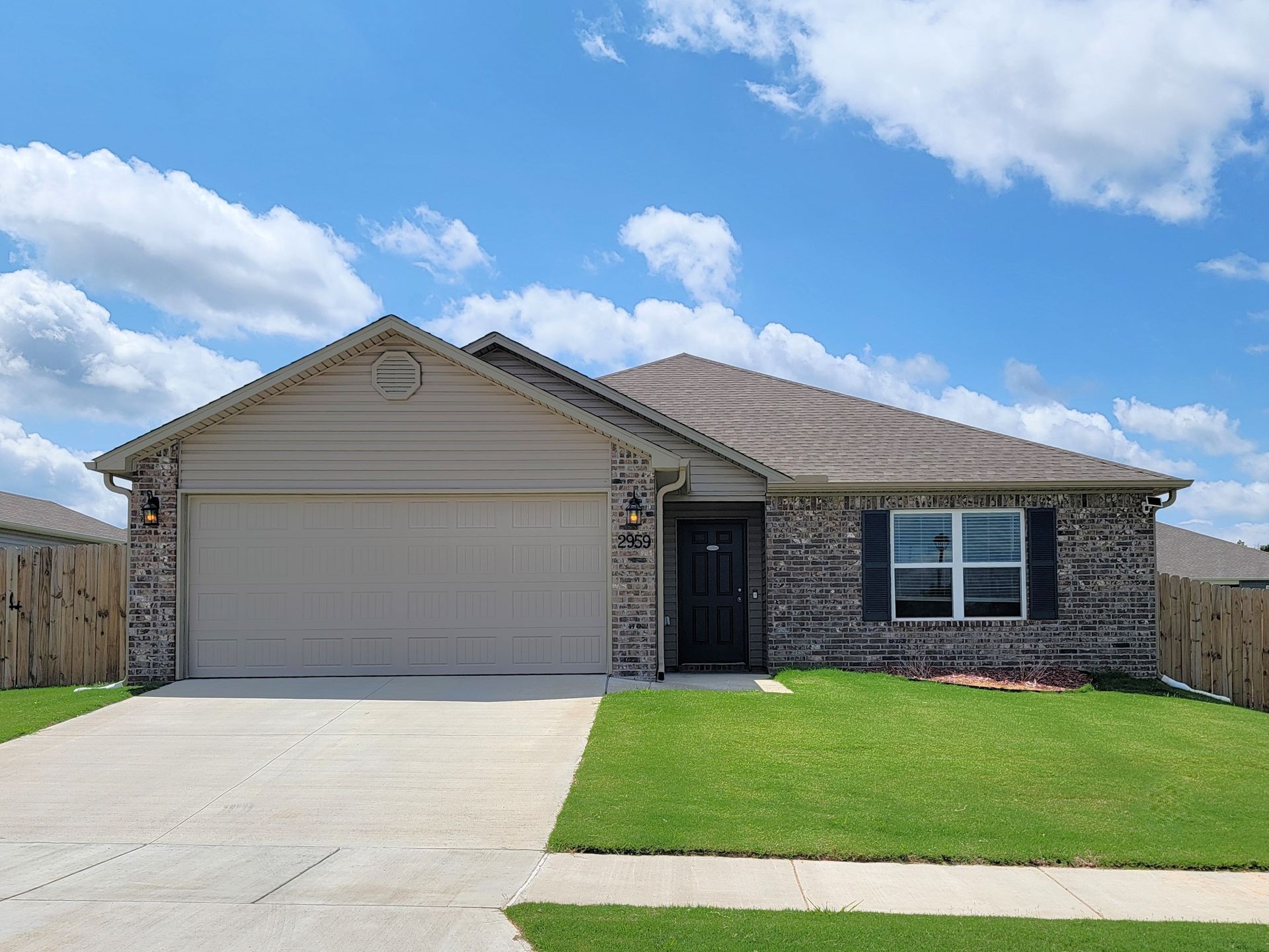 Single-story beige house with stone trim, two-car garage, driveway, and green lawn under a blue sky