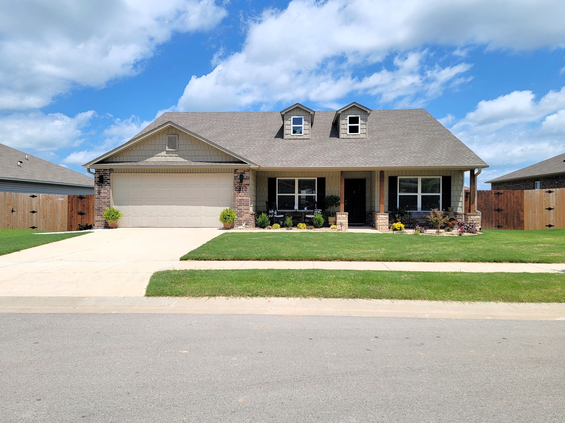 Single-story suburban house with a two-car garage, front porch, and a wide driveway under a blue sky